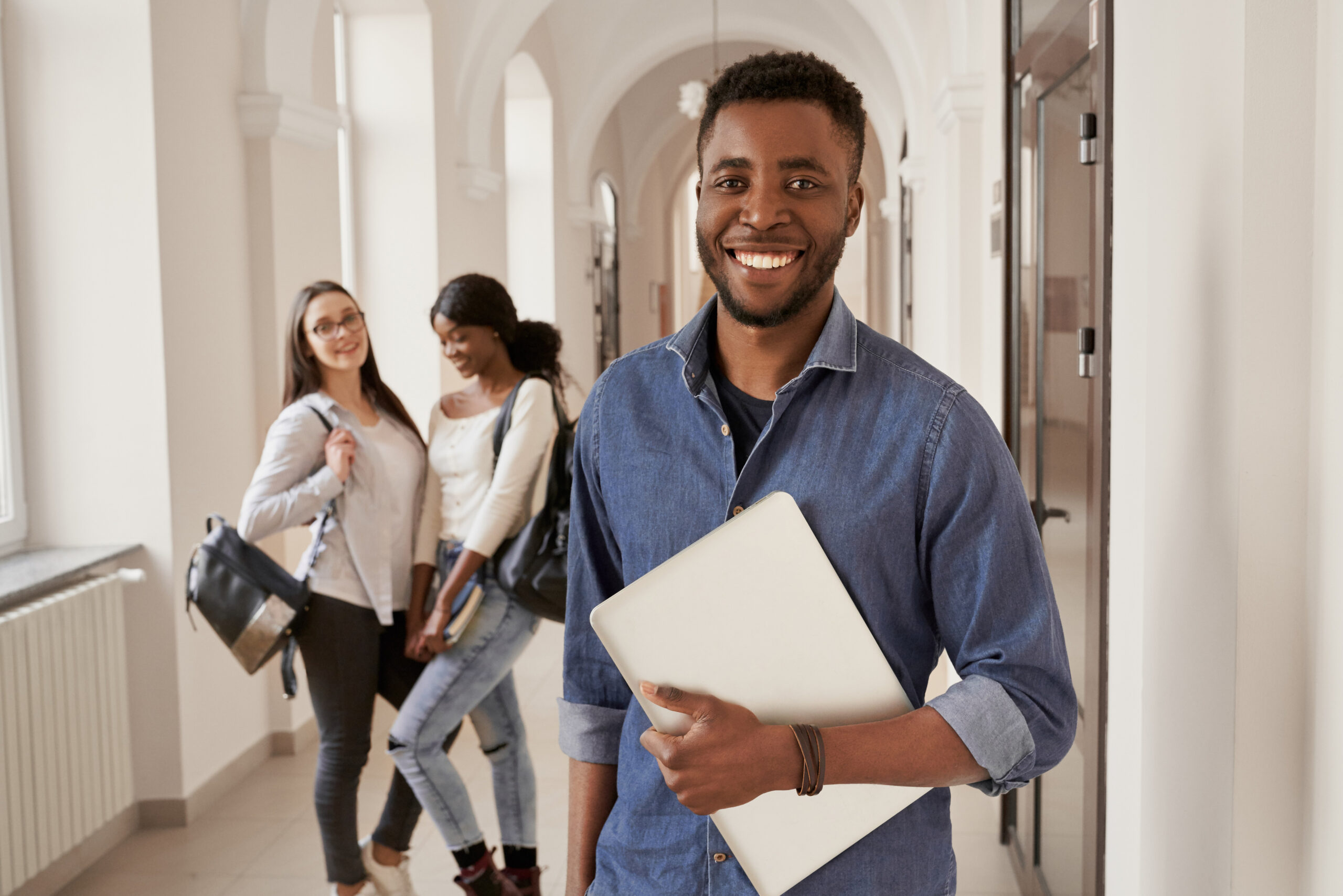 Positivity African student wearing shirt holding notes, going to lecture and smiling. Happy male studying at international university. Two pretty girls with backpacks on background.